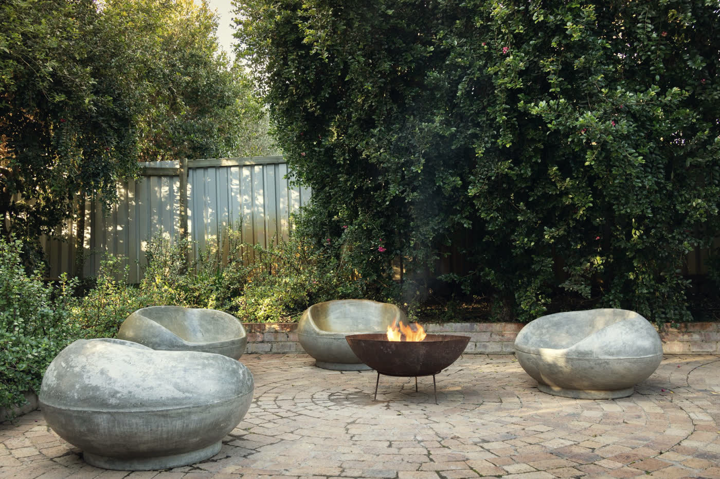 The same garden corner with a circular tumbled-cobble patio laid in a radial fan, a lit steel fire bowl at its centre and three cast-concrete boulder seats around it.