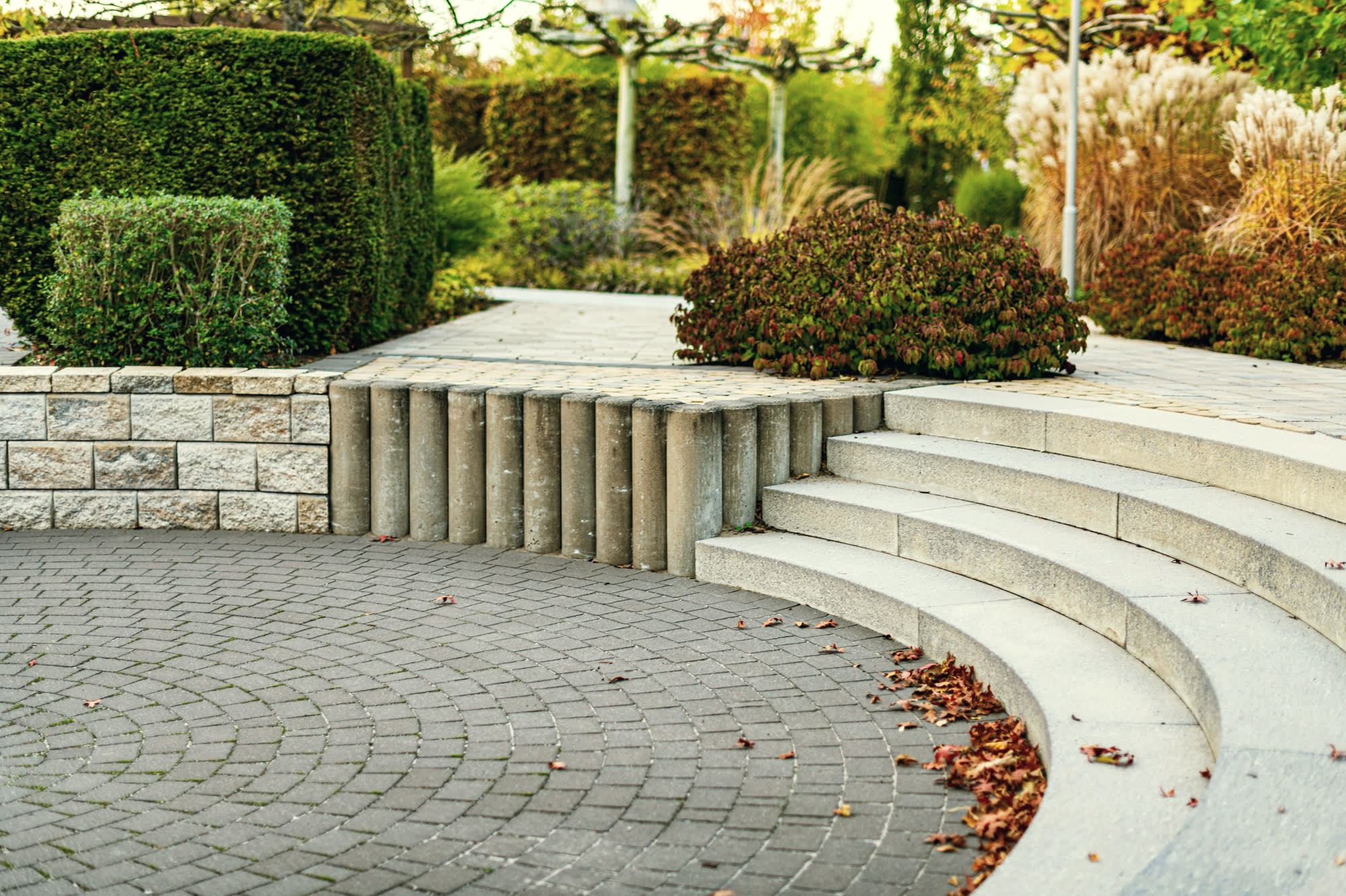 Curved sawn-granite steps sweeping into a circular fan-pattern paver terrace, beside a split-face block retaining wall, with clipped hedging and autumn grasses behind.