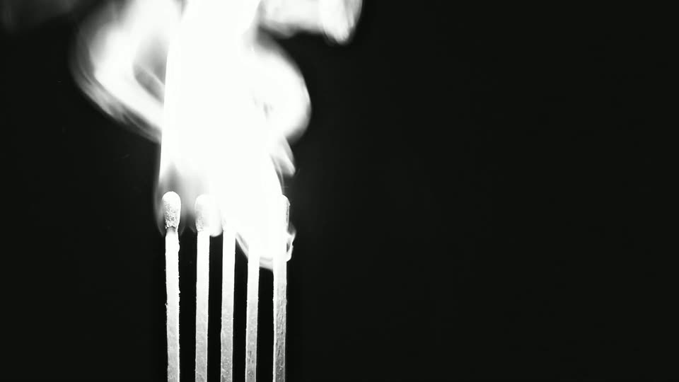 Frame 008, the flare: a tall white flame plume engulfs the row of match heads and fills the upper left of the frame, its top turning over into smoke. The five sticks below stand lit against black.